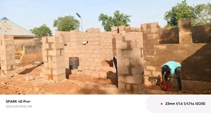 A longer run of block walls across the site, with a man in a turquoise shirt bending over a red bucket at the right and another worker at the far left.
