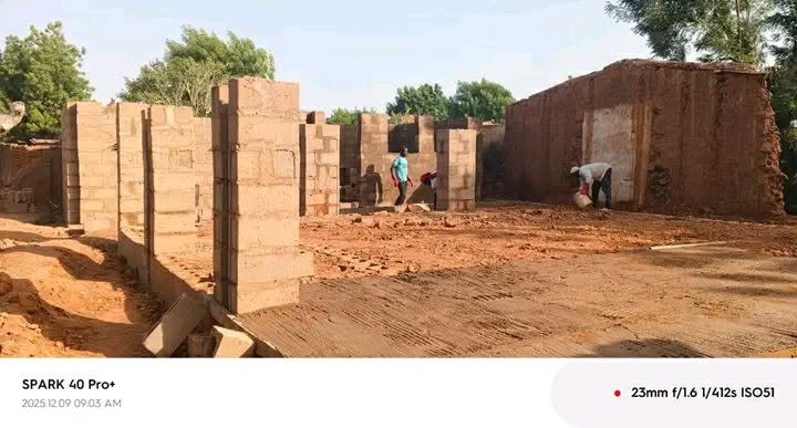 Several separate block wall panels standing in a sandy compound, with two men working between them and an older rendered building with a doorway at the right.
