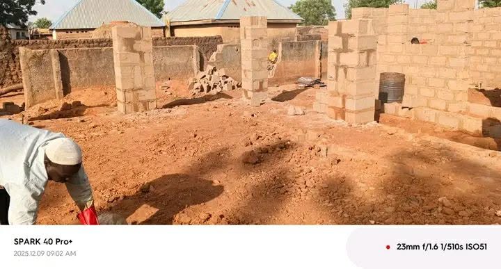 A man in a cap bending over a red bucket at the edge of the site, with a pile of rubble, more block walls and a basin set into the wall behind him.