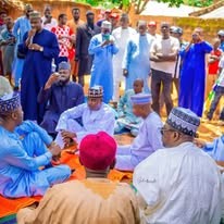 A large gathering of men in white and blue robes seated on blue and orange mats beside an earthen building.