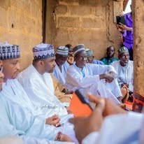Men in white robes and patterned caps seated in rows at a gathering, with a hand holding a phone in the foreground.