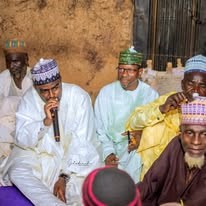 Men seated on mats at a community meeting beside an earthen wall, one speaking into a microphone.