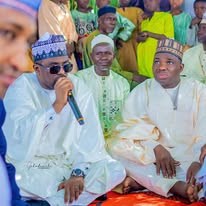 Hon. Umar Yusuf Yabo speaking into a handheld microphone outdoors, wearing sunglasses and a patterned cap, surrounded by standing men.