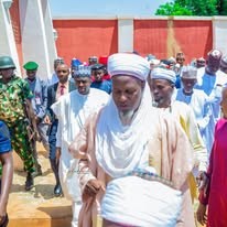 Hon. Umar Yusuf Yabo seated in a white turban and cream robe, with a crowd standing behind a red and white painted wall.