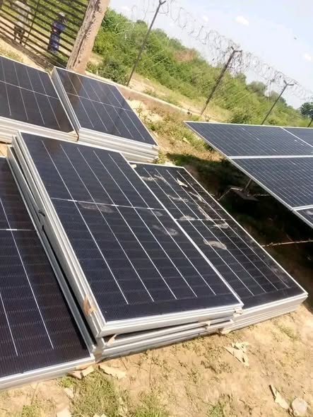 Photovoltaic panels stacked on cleared ground inside a compound, with a wire fence and thorny scrub beyond.