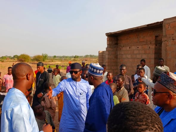 Hon. Umar Yusuf Yabo, in a light blue outfit and sunglasses, in conversation with community members beside a partly built block wall.