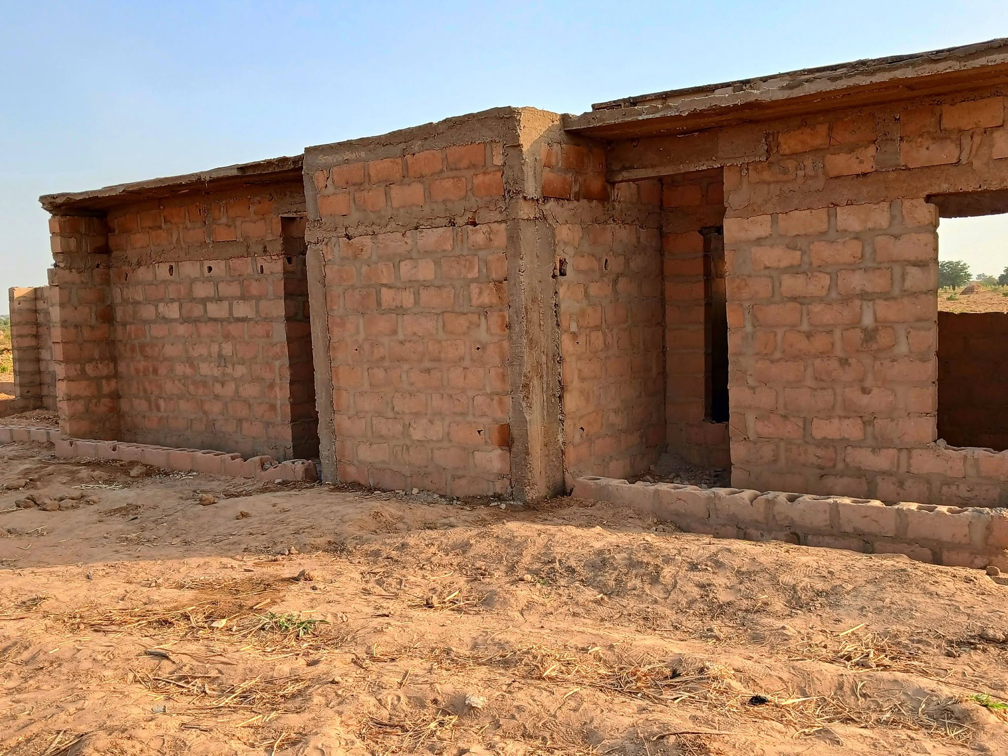 A long single-storey building under construction in unfired block, its door and window openings still empty and the walls uncapped.