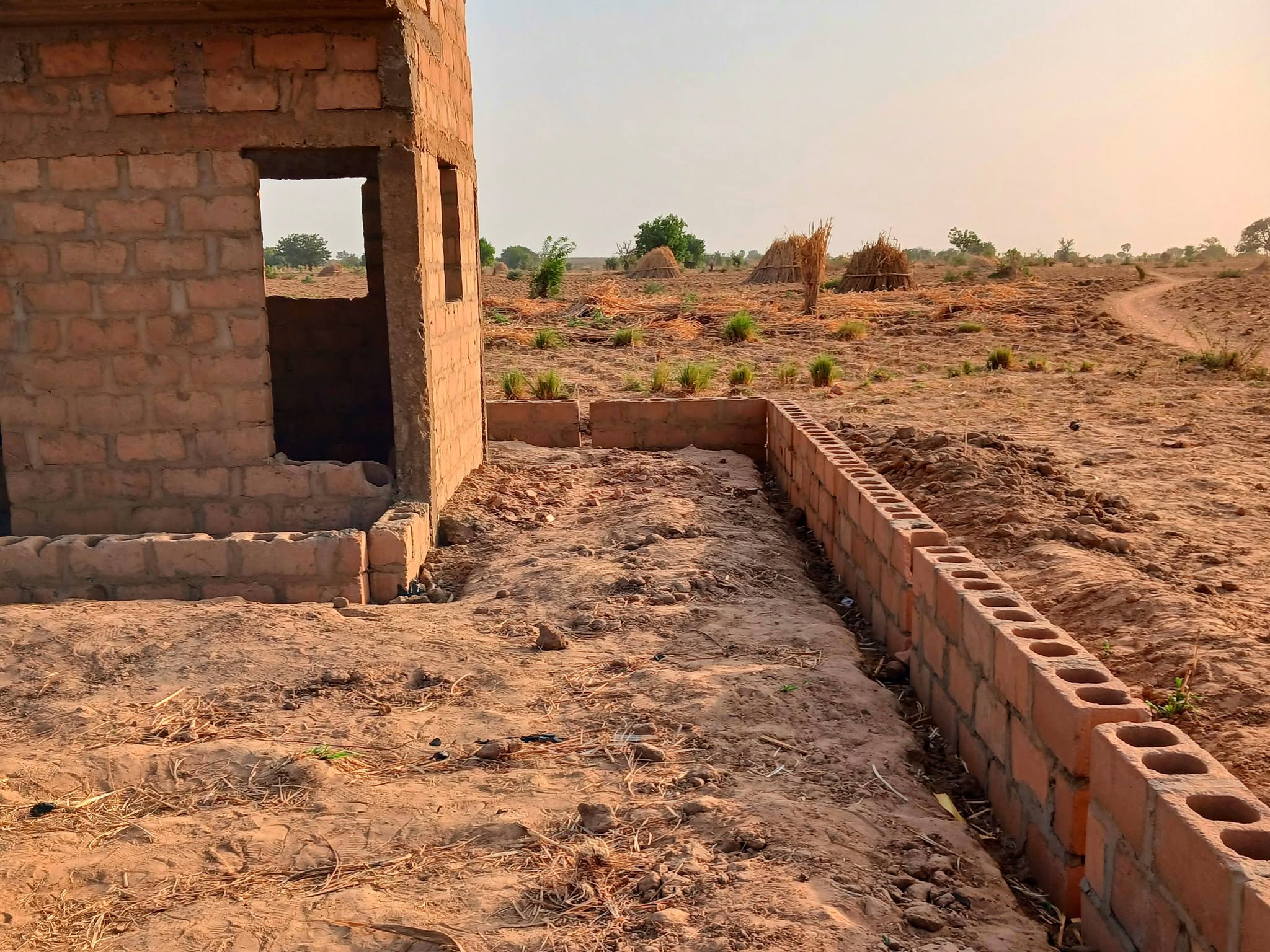 The corner of the same block building, with a bare doorway and fresh blockwork running out across the sandy, harvested ground.
