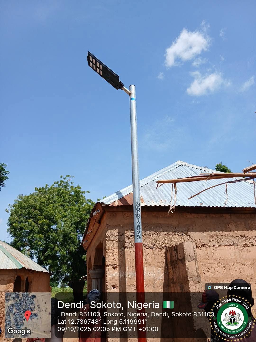 A new solar street light: a slim red-and-white pole with a rectangular luminaire, raised against the sky above a mud-walled house.