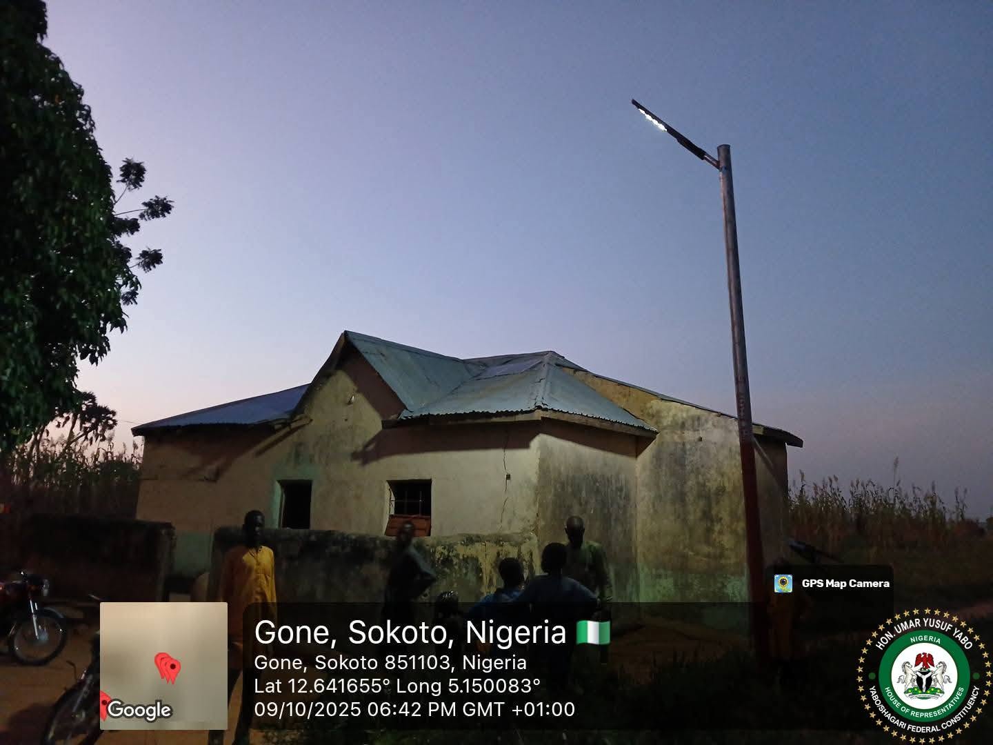 A solar street light at dusk beside a cream house, its luminaire lit, with residents standing on the road below.