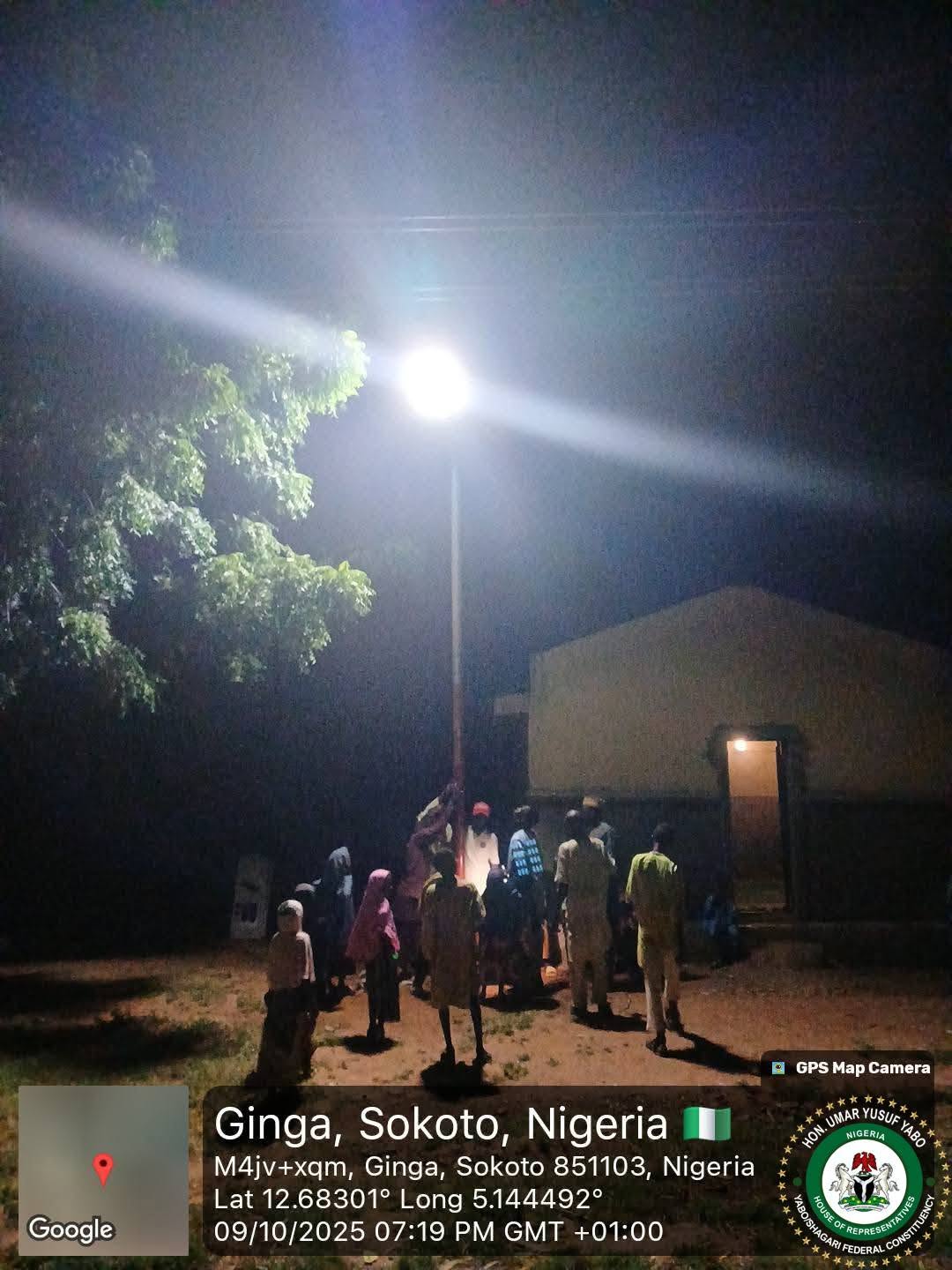 A brighter night view of the same lit street light, throwing a broad beam across the branches of a nearby tree, with a crowd gathered below.