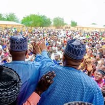 A large outdoor rally crowd seen from behind two men in caps, one with a hand raised, with trees and the gathering stretching into the distance.