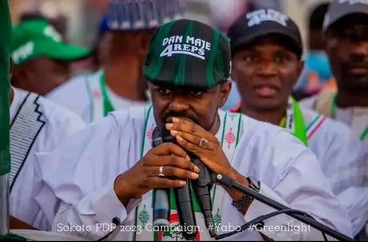 Hon. Umar Yusuf Yabo speaking into a microphone at a campaign event, wearing a green and black cap marked “Dan Maje 4Reps” and a white robe with green and pink embroidery.