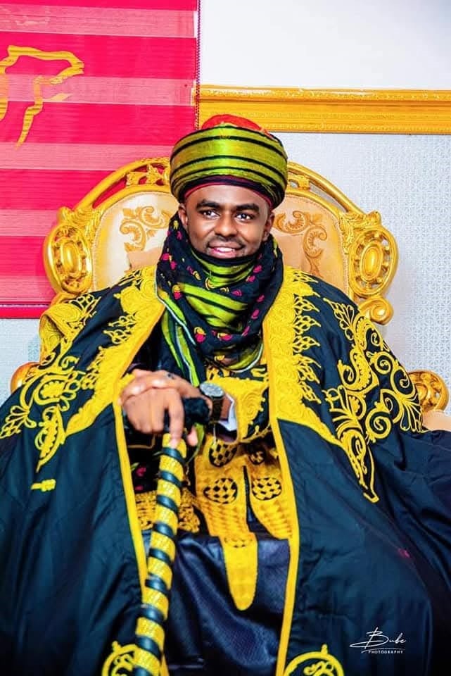 Hon. Umar Yusuf Yabo in traditional regalia, seated on an ornate carved chair and holding a ceremonial sword.