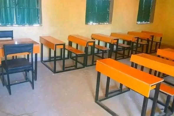 Inside a classroom, rows of orange-topped single benches with black metal frames, set on a clean concrete floor beneath green barred windows.