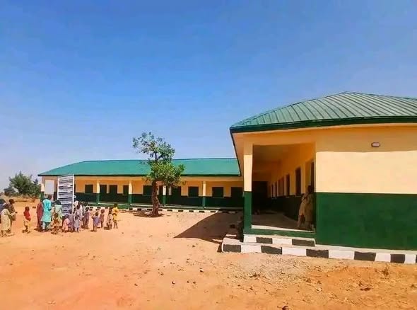 A wider view of the campus, showing two cream and green blocks with green roofs across a sandy courtyard, with a tree planted in the middle and a group of pupils gathered to the left.
