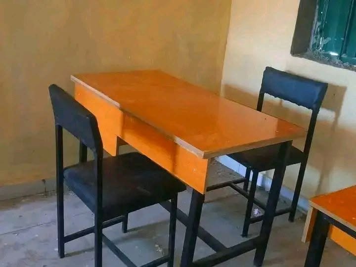 A close view of a single orange-topped desk and two black plastic chairs in a corner of the classroom, beside a green barred window.