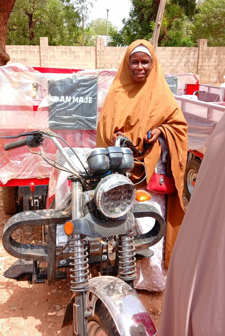 Two women at the handover, one in pink and one in cream, beside a motorcycle and a wrapped “DAN MAJE” tricycle box.