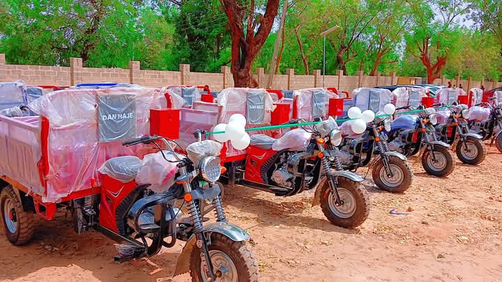 Rows of new motorcycles and tricycle cargo boxes branded “DAN MAJE”, wrapped in protective plastic, lined up on cleared ground at the December 2024 distribution.