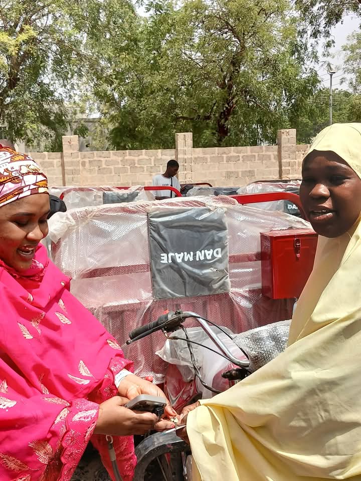 A woman in a mustard shawl seated on a newly distributed motorcycle, one hand on the handlebar, with the wrapped tricycle boxes behind her.