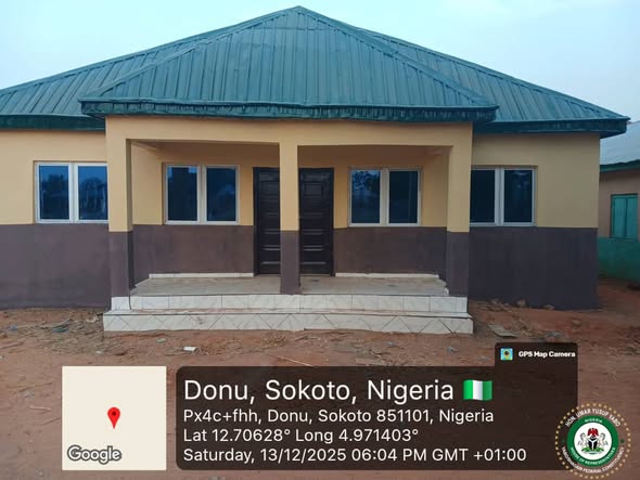 Front elevation of the renovated Donu Primary Healthcare Centre: a single-storey cream building with a dark brown lower band, a green hipped metal roof, white-framed windows and black double doors reached by tiled steps.