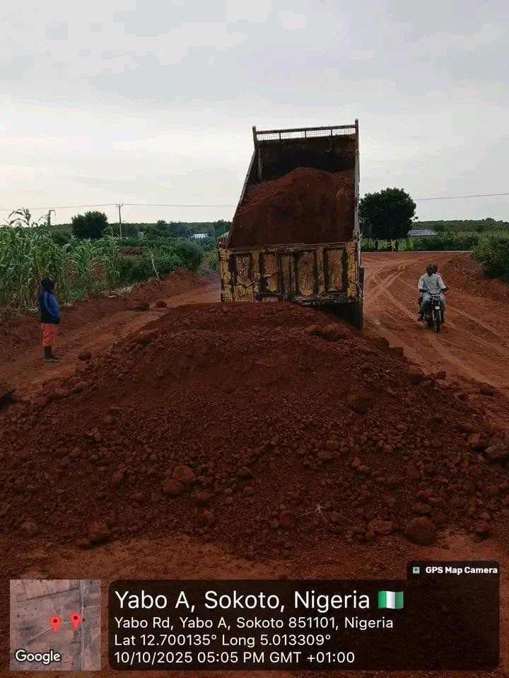 A tipper truck standing with its bed raised, dropping a mound of red laterite onto the road, a man watching from the verge and a motorcyclist passing on the dirt track beside it.