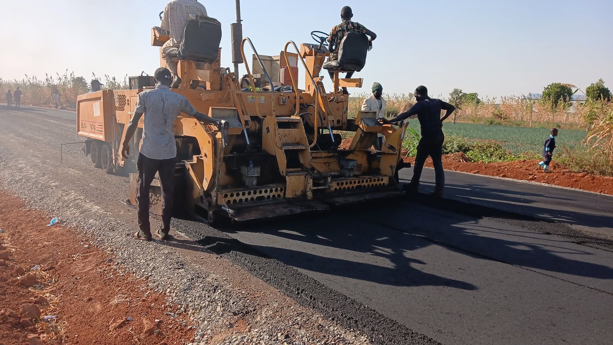 A yellow paving machine laying hot bitumen across the carriageway, with crew members walking behind it and one seated at the controls.