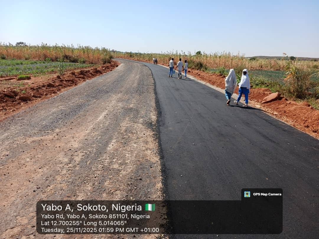 The finished tarred road curving away between maize fields, with a gravel shoulder and schoolchildren in white and blue uniforms walking along it.