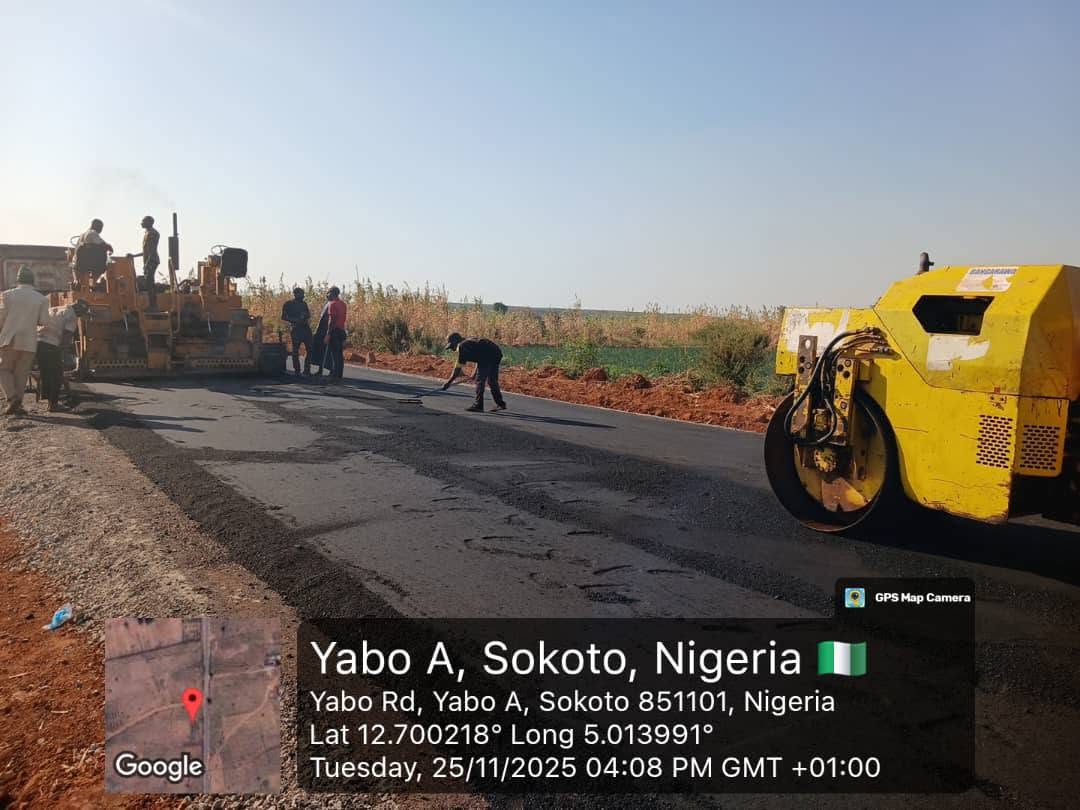A yellow road roller working over the fresh black bitumen surface while the paving machine and its crew wait further back.
