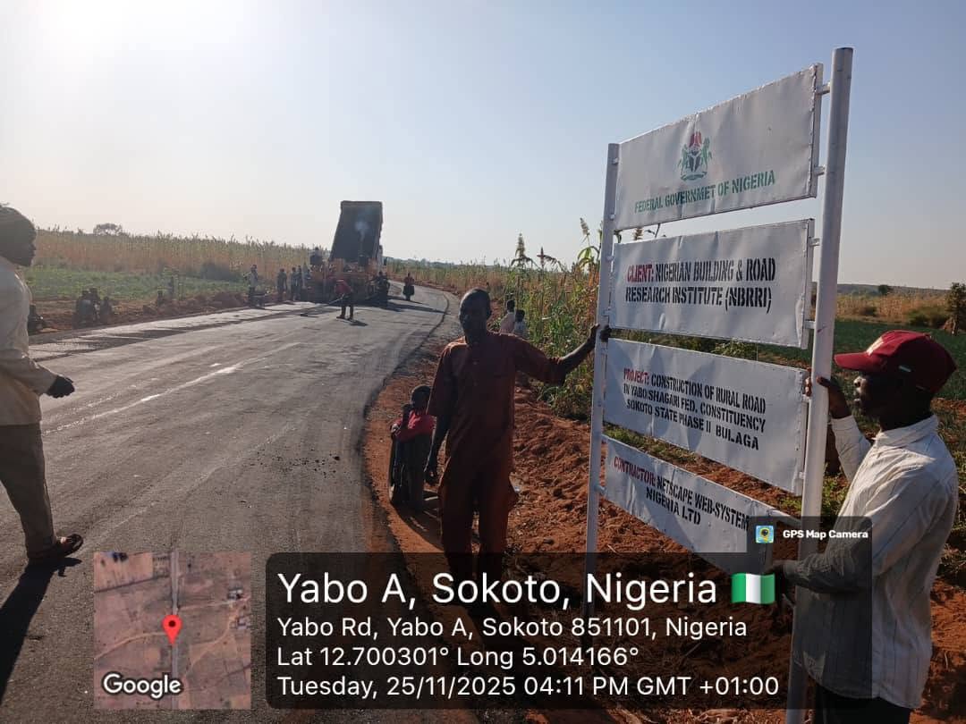 Men holding up the Federal Government of Nigeria project signboard for the Yabo Shagari rural road, phase two at Bulaga, with the newly laid road and a truck behind it.